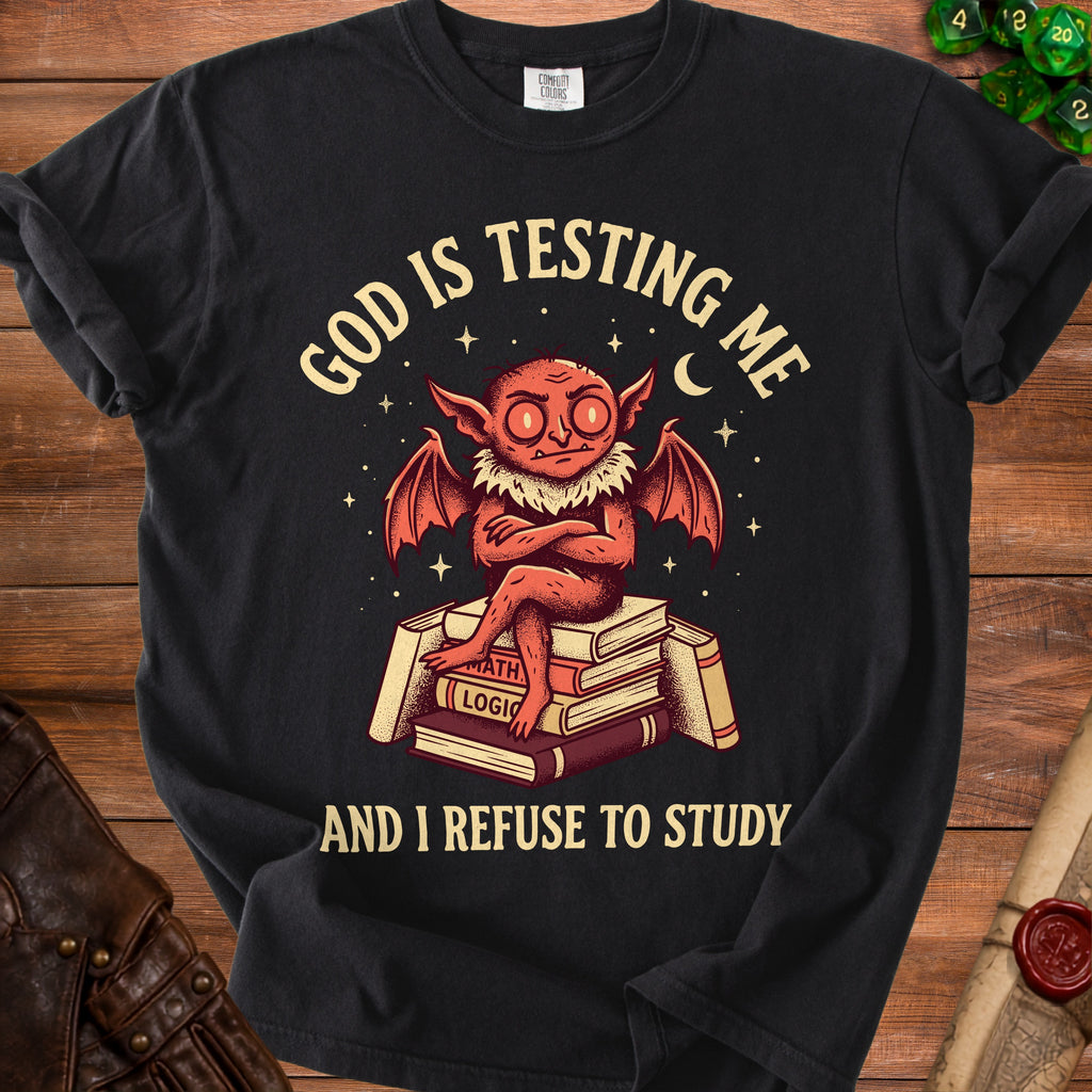Black t-shirt with a cartoon demon sitting on books with text "God is testing me and I refuse to study".
