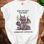 White t-shirt with a pink dragon wearing sunglasses, holding a coffee mug. Text reads "Hop up out the bed summon my coffee on".