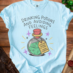 Light blue t-shirt with "DRINKING POTIONS AND AVOIDING FEELINGS" and a potion tag.