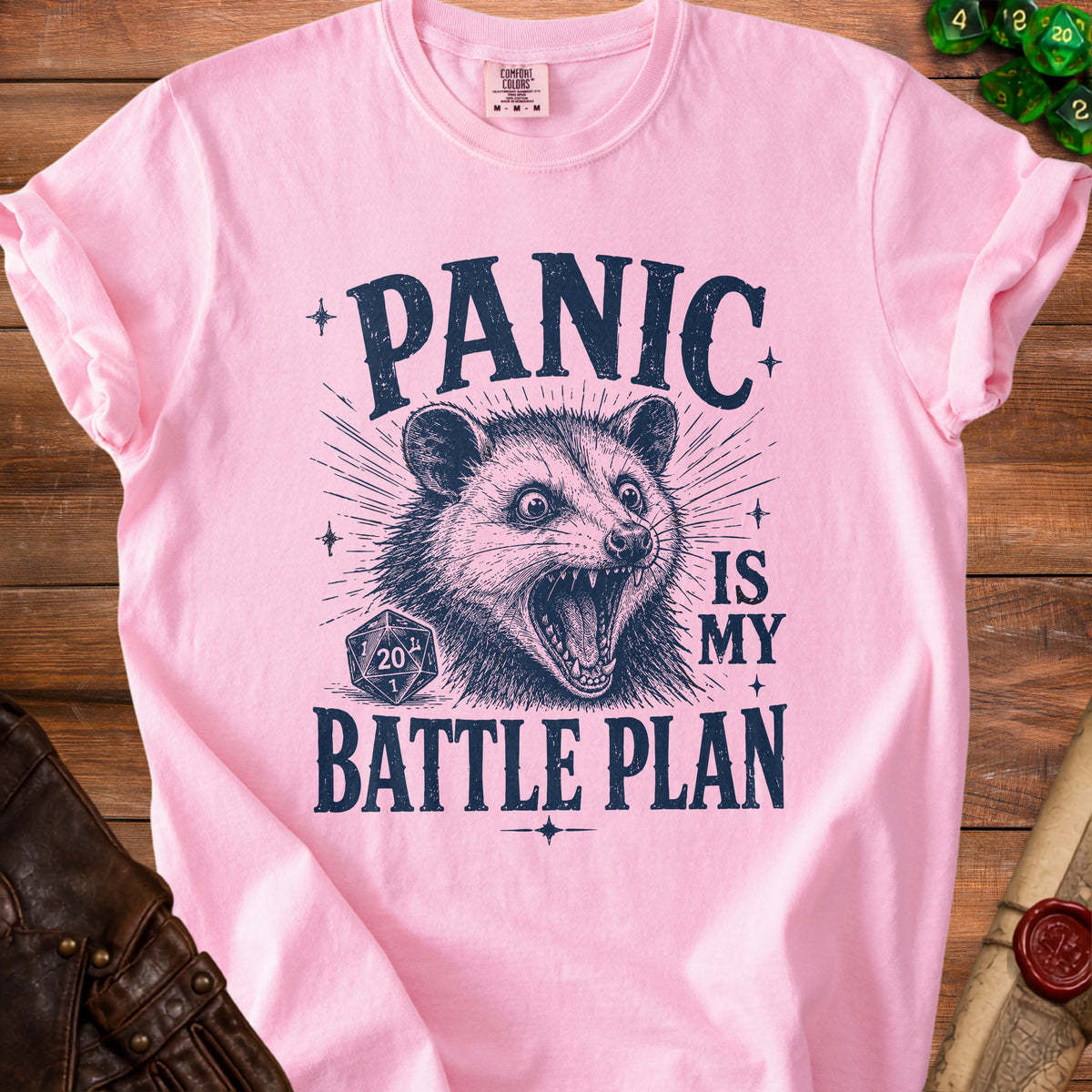 Pink t-shirt with dark blue text and graphic of a panic-stricken opossum and dice.