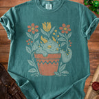 Teal t-shirt with a sleeping dragon in a potted plant surrounded by flowers.