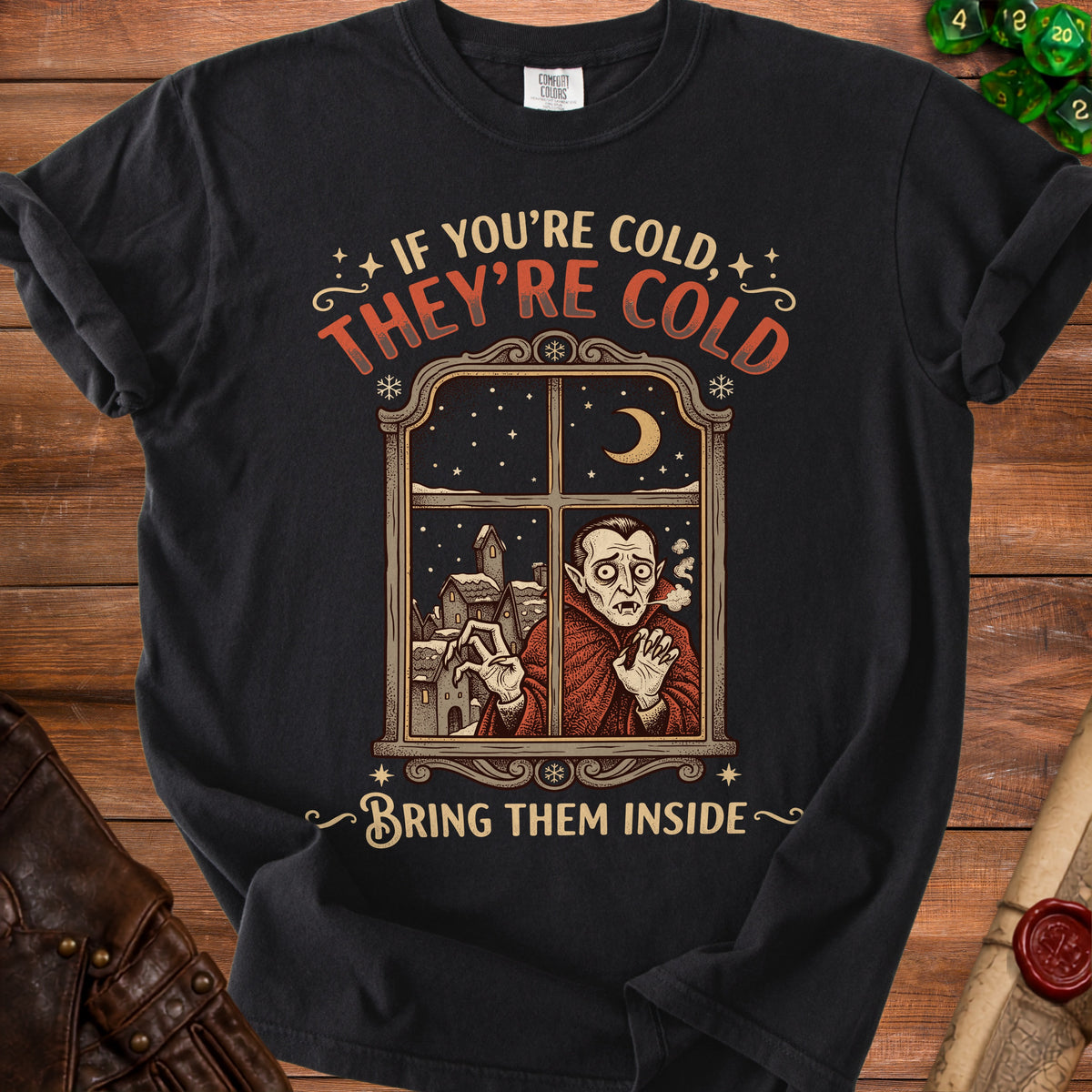 T-shirt with vampire looking out a window in a snowy village saying "If you're cold, they're cold. Bring them inside."