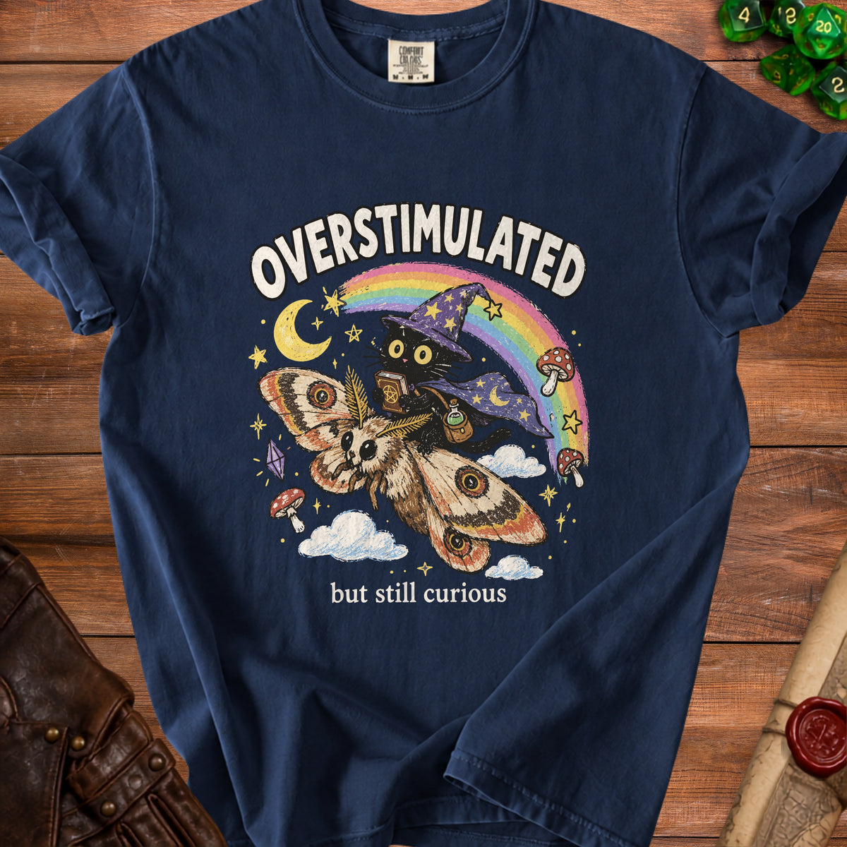 Navy t-shirt with graphic of a moth, black cat witch, rainbow, and mushrooms with text "Overstimulated but still curious".