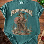 "Frontier Mage" graphic on a teal t-shirt featuring a witch in a western landscape.