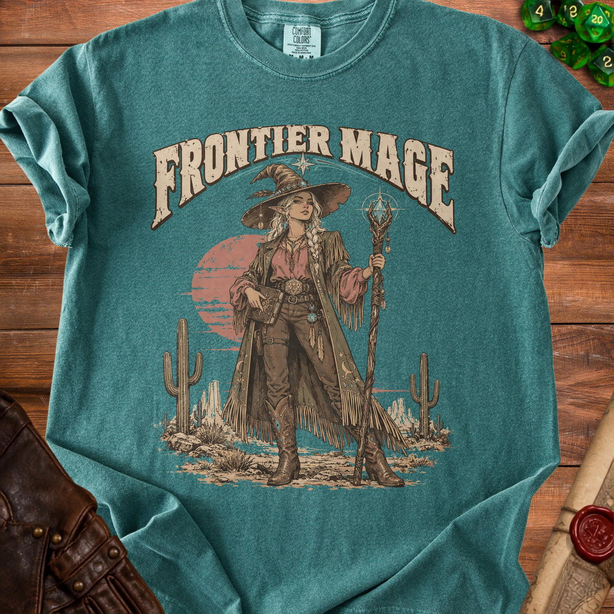 "Frontier Mage" graphic on a teal t-shirt featuring a witch in a western landscape.