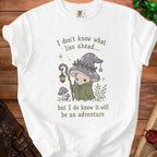 White t-shirt with a mushroom character wearing a hat and cloak, holding a staff, and text about adventure.