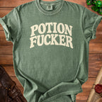 Green t-shirt with "POTION FUCKER" text in beige, placed on a wooden surface.