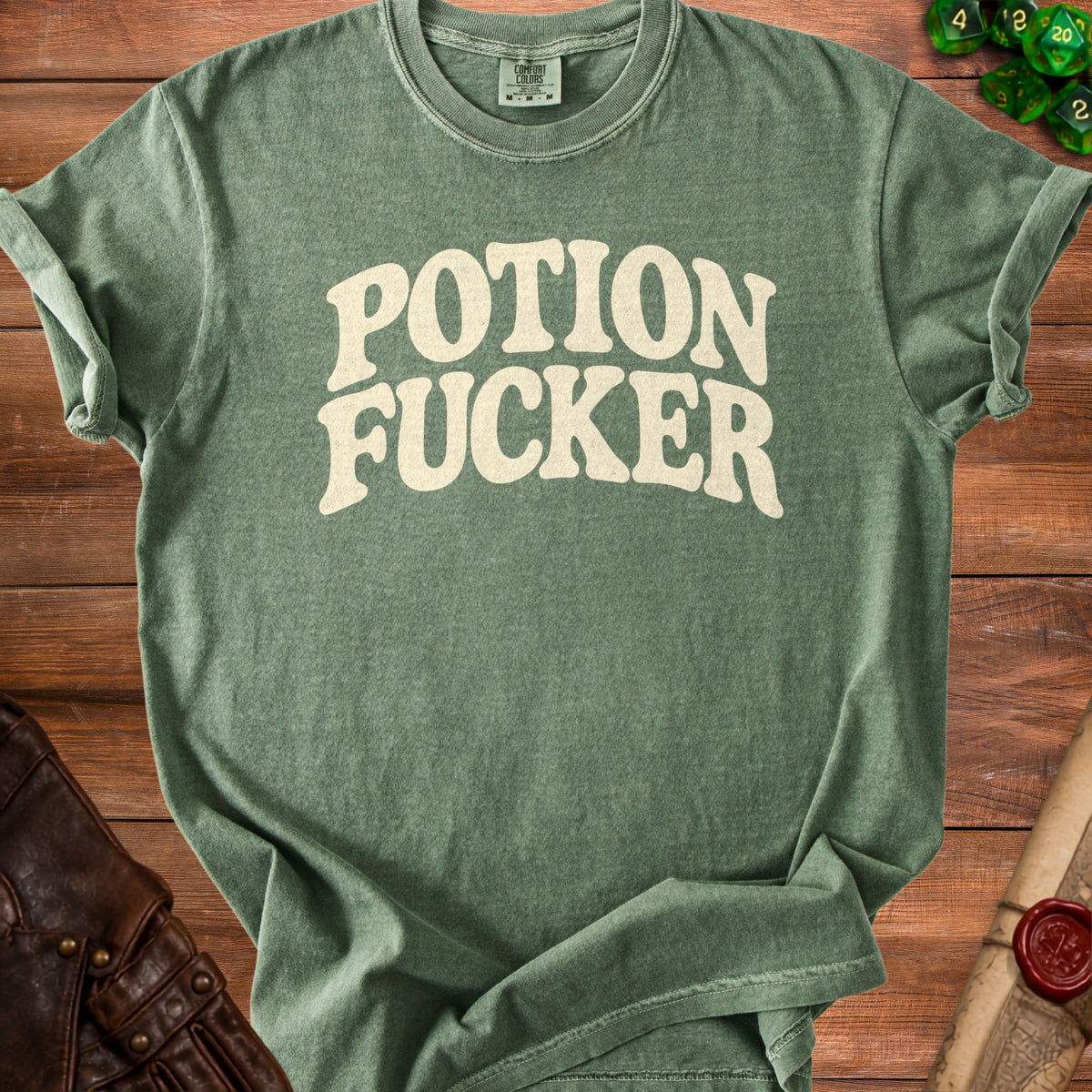 Green t-shirt with "POTION FUCKER" text in beige, placed on a wooden surface.