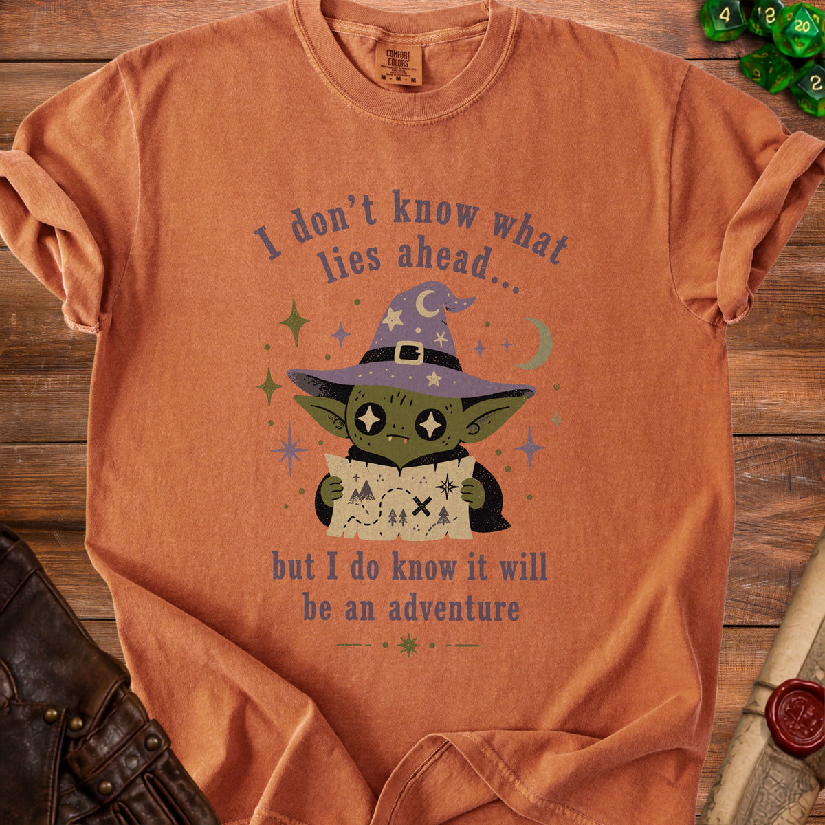 Orange t-shirt with a green goblin in a witch hat holding a map, text reads "I don't know what lies ahead...but I do know it will be an adventure".