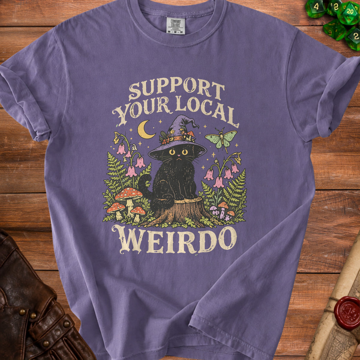 A purple crewneck t-shirt featuring a graphic of a black cat in a witch hat, surrounded by mushrooms and the text "SUPPORT YOUR LOCAL WEIRDO."