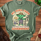 Green t-shirt graphic with sad goblin farmer, barn, windmill, and text "MY AURA FARM was Shut Down by the GOVERNMENT."