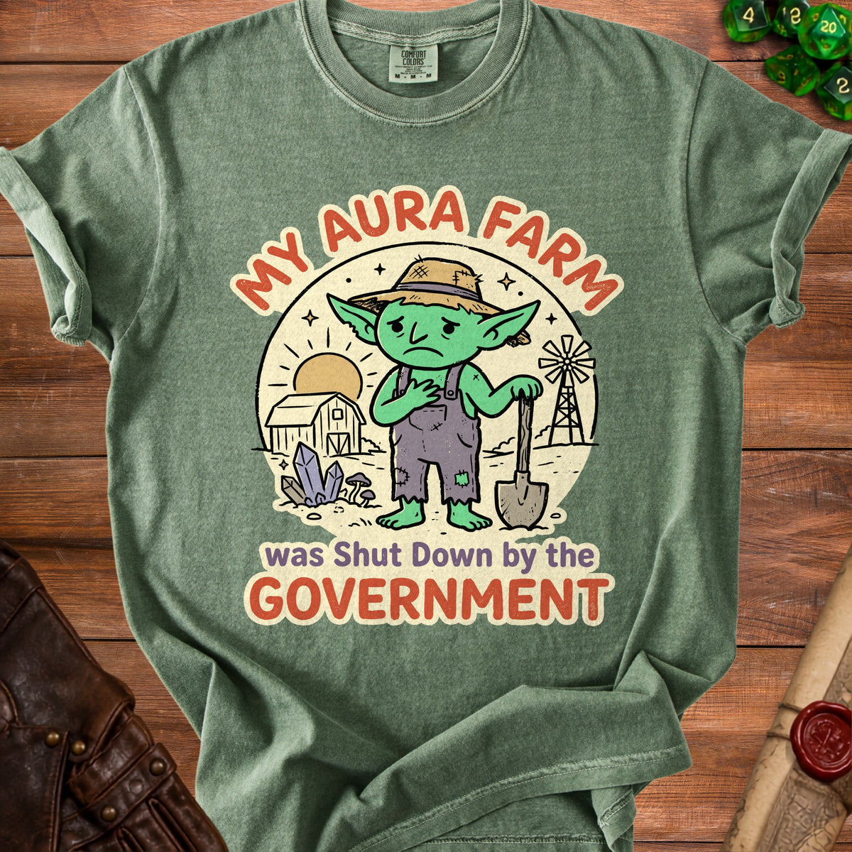 Green t-shirt graphic with sad goblin farmer, barn, windmill, and text "MY AURA FARM was Shut Down by the GOVERNMENT."