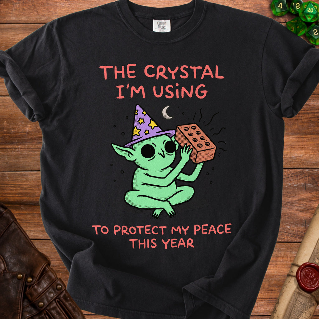 Black t-shirt with green creature wearing wizard hat, holding brick. Text: "The crystal I'm using to protect my peace this year".