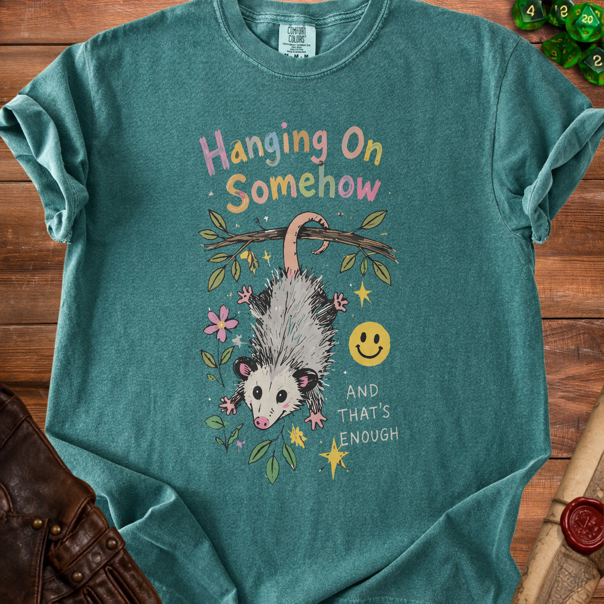 Teal shirt with opossum hanging from branch, text "Hanging On Somehow AND THAT'S ENOUGH".