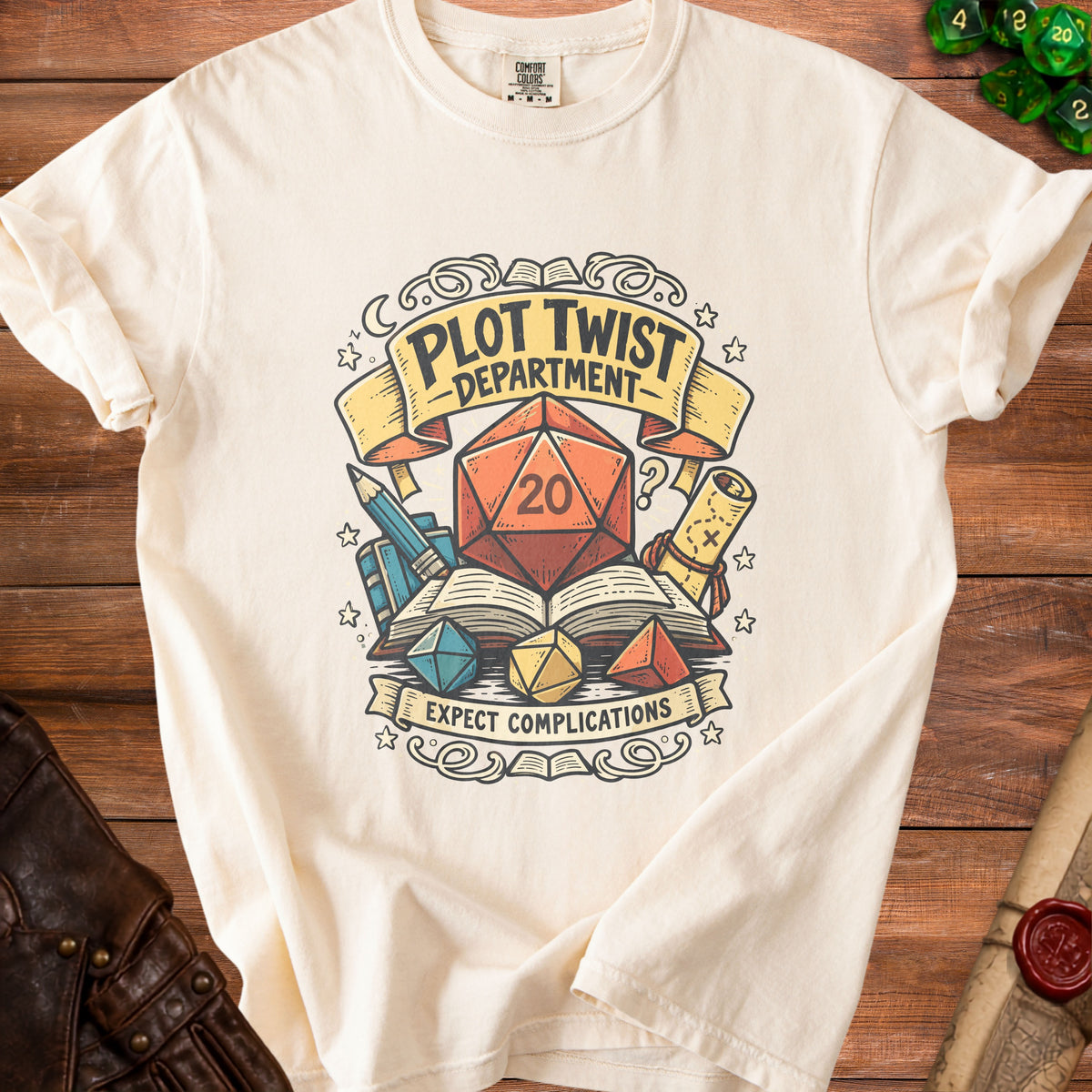 Cream t-shirt with a d20 die, books, and banners saying "Plot Twist Department" and "Expect Complications".