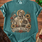 Skeleton in robes holding potion and book, surrounded by flowers, "Still Healing" text.