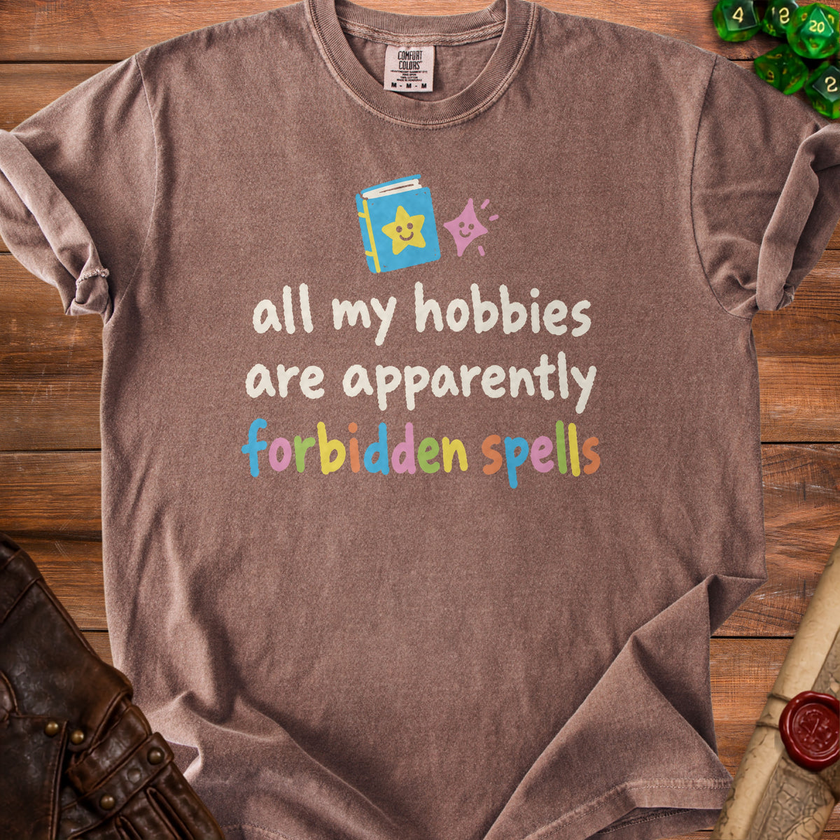 Brown t-shirt with "all my hobbies are apparently forbidden spells" text and cartoon book graphic.