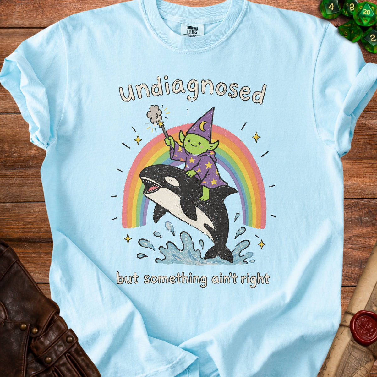 Light blue t-shirt with cartoon of wizard riding orca over rainbow, text "undiagnosed but something ain't right".