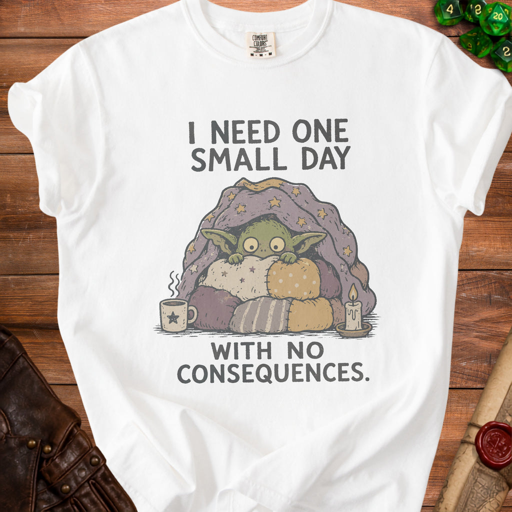 White t-shirt with a green creature under a starry blanket, text says "I need one small day with no consequences."