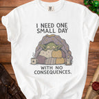 White t-shirt with a green creature under a starry blanket, text says "I need one small day with no consequences."