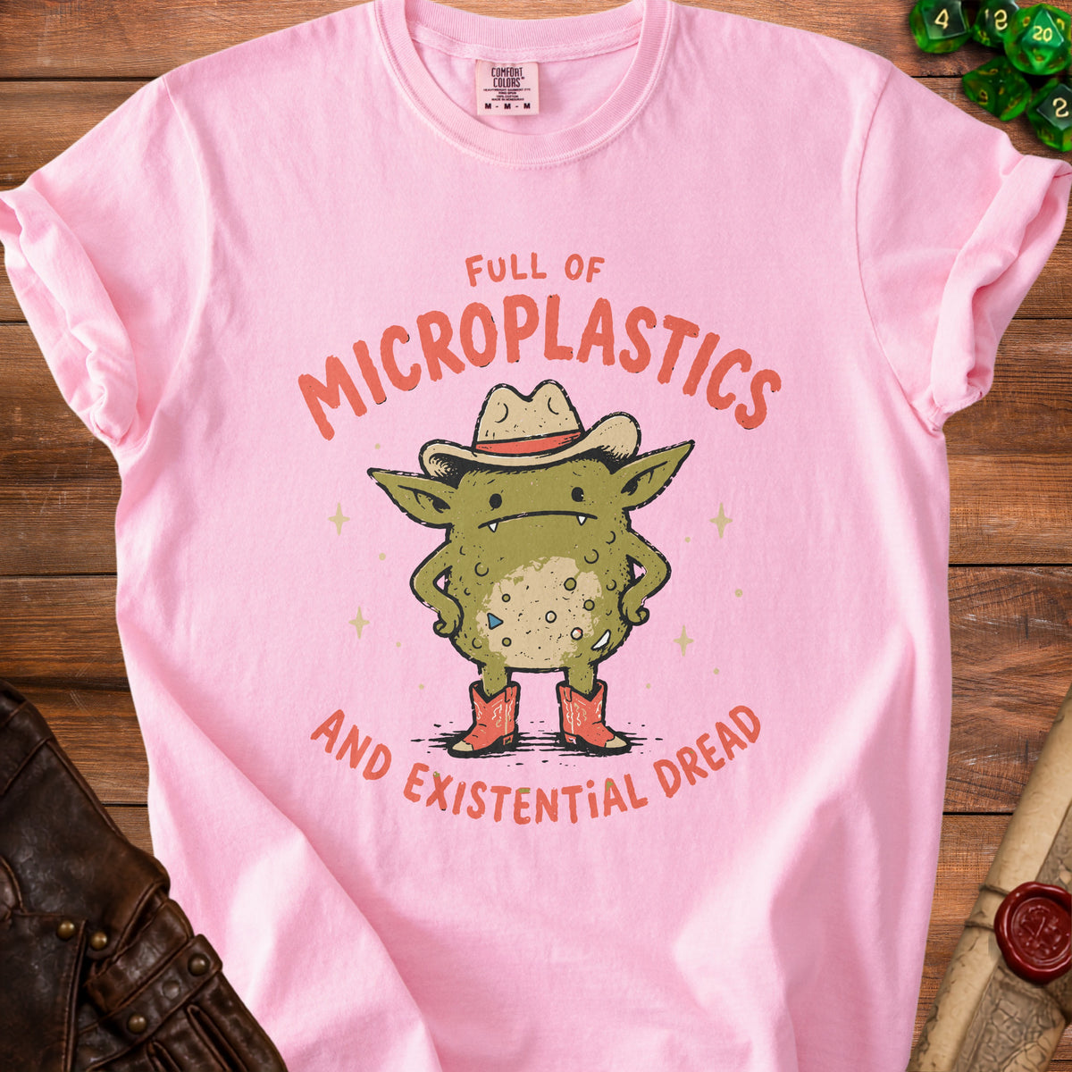 Pink t-shirt with a green monster wearing a cowboy hat and boots, text "FULL OF MICROPLASTICS AND EXISTENTIAL DREAD".