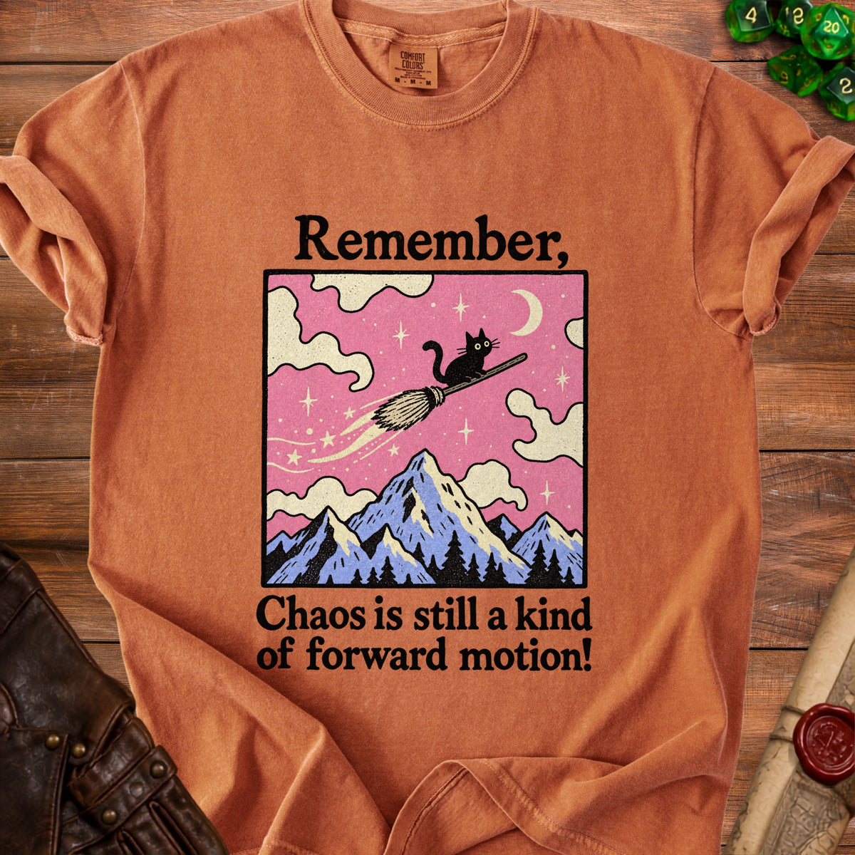 Orange t-shirt with black text and a graphic of a cat on a broom flying over mountains.