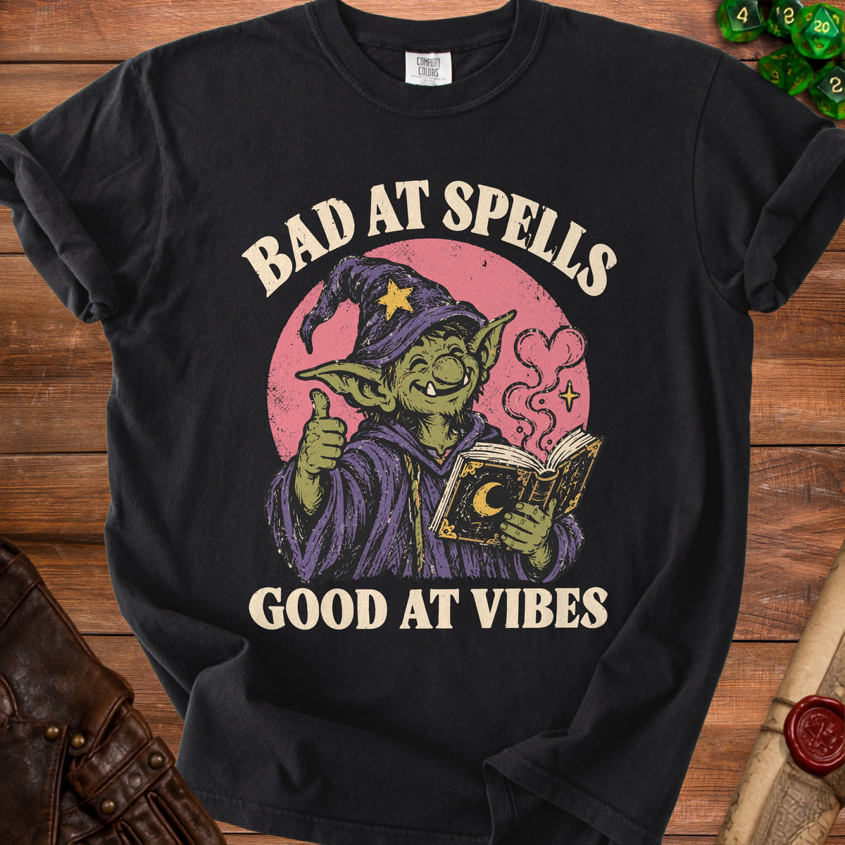 Black t-shirt with graphic of a green goblin reading a book, text "BAD AT SPELLS GOOD AT VIBES".