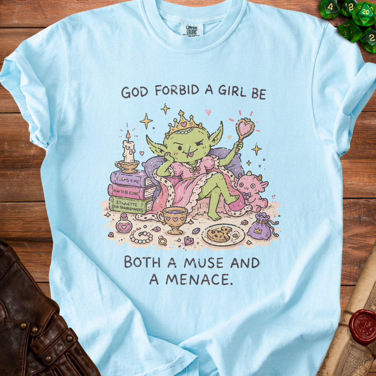Light blue t-shirt with a cartoon goblin queen and text "GOD FORBID A GIRL BE BOTH A MUSE AND A MENACE."