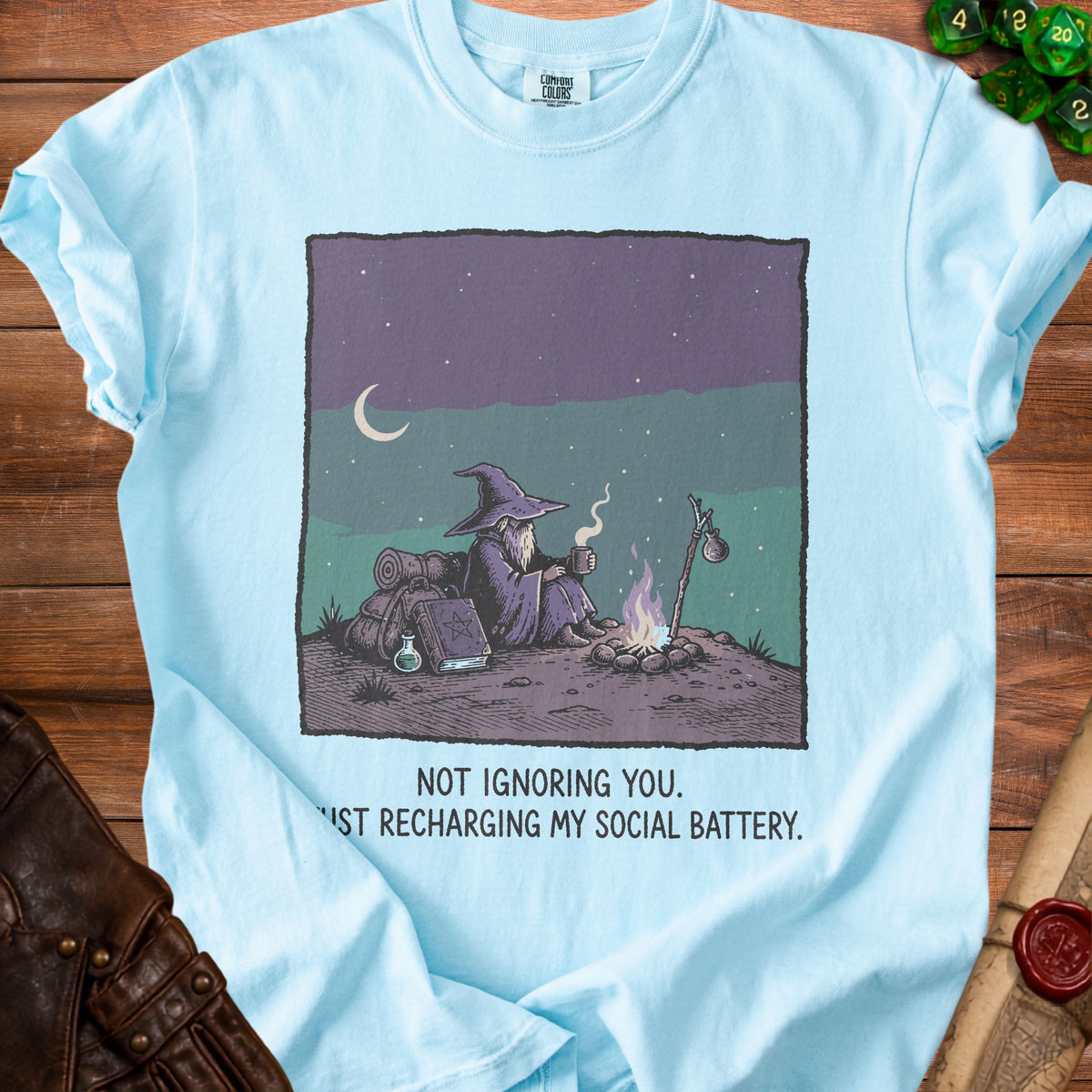 Light blue t-shirt with wizard sitting by campfire and text "NOT IGNORING YOU. JUST RECHARGING MY SOCIAL BATTERY."