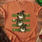 Orange t-shirt with three cartoon frogs wearing witch hats on green stripes.