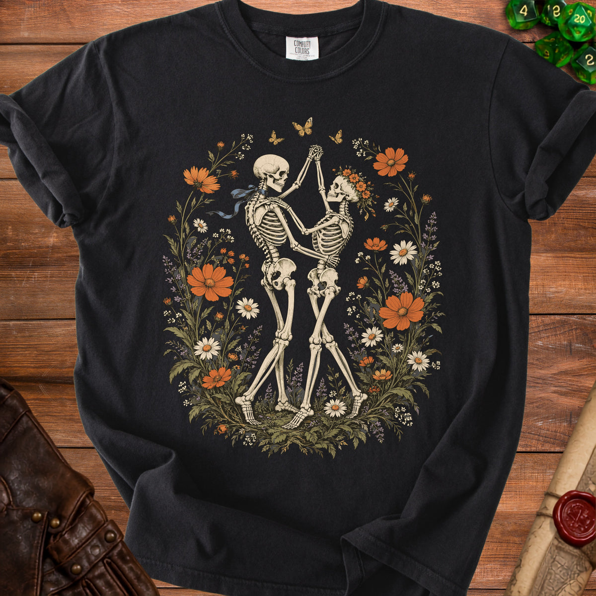 A black t-shirt features a vintage illustration of two skeletons dancing amidst a floral wreath.