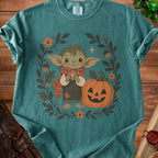 Green t-shirt with illustration of cute creature, jack-o'-lantern, and autumn leaves.