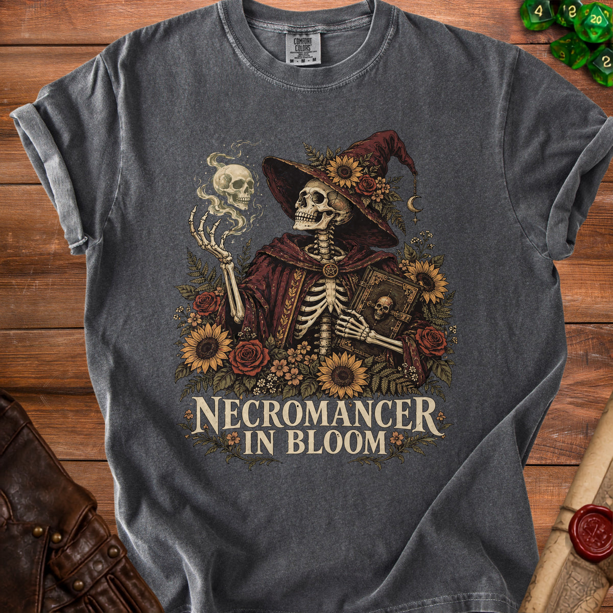 A dark gray t-shirt featuring a skeletal wizard surrounded by flowers, holding a book, and casting magic, with the text "Necromancer in Bloom."