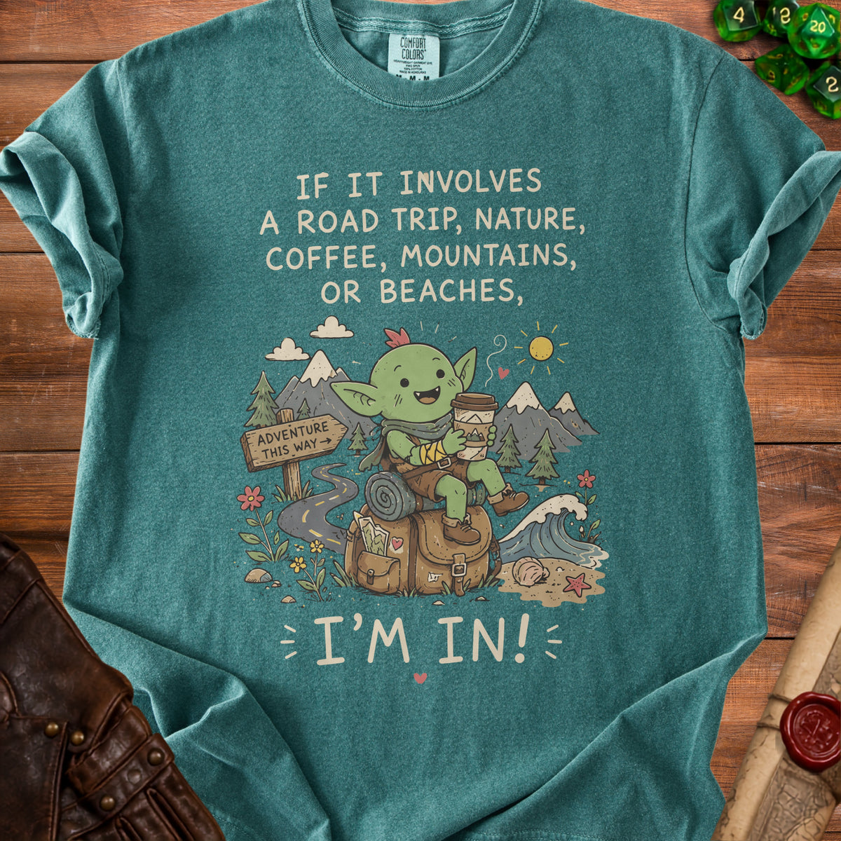 Green t-shirt with a cartoon goblin drinking coffee and text "IF IT INVOLVES A ROAD TRIP, NATURE, COFFEE, MOUNTAINS, OR BEACHES, I'M IN!".