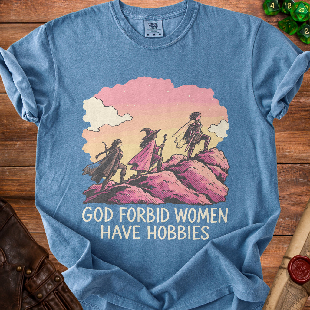Blue t-shirt featuring an illustration of three female adventurers on a mountain with text reading GOD FORBID WOMEN HAVE HOBBIES.
