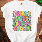White t-shirt with "Stay Weird Little Goblin" text, mushrooms, goblins, and stars.