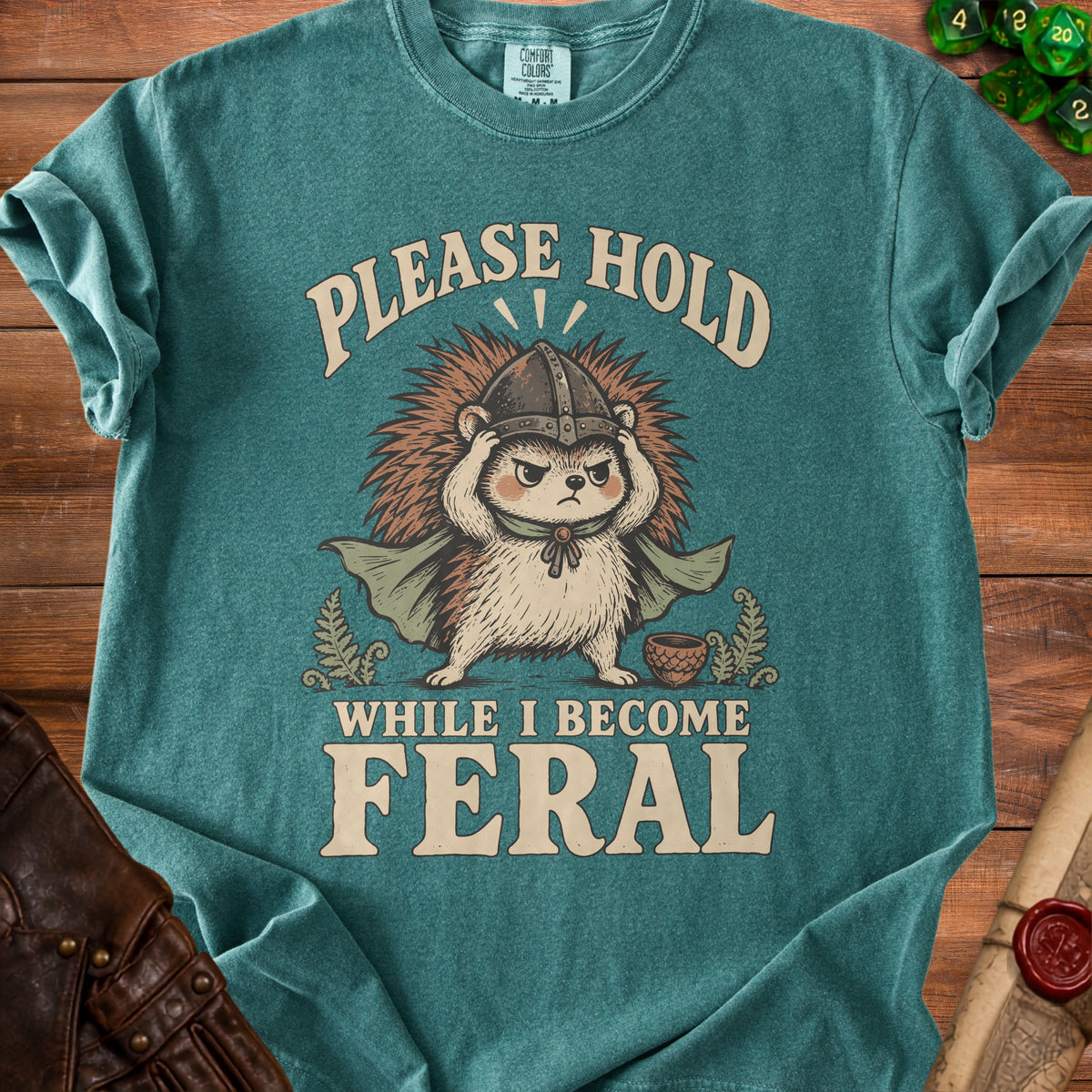Teal t-shirt with a grumpy hedgehog wearing a helmet and cape, text "PLEASE HOLD WHILE I BECOME FERAL".