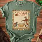 Green t-shirt featuring a medieval-style illustration of a cook and dragon beside a bowl, with text "A Modest Request".