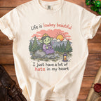 Cream t-shirt with cartoon goblin on rock, sunset, and text "Life is lowkey beautiful I just have a lot of hate in my heart".