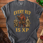 Dark gray t-shirt with a Comfort Colors tag featuring a dragon, dwarf, and wizard lifting weights with text "EVERY REP IS XP".