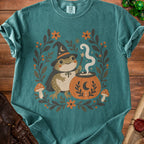 Frog wizard holding wand next to cauldron on teal t-shirt with floral and mushroom border.