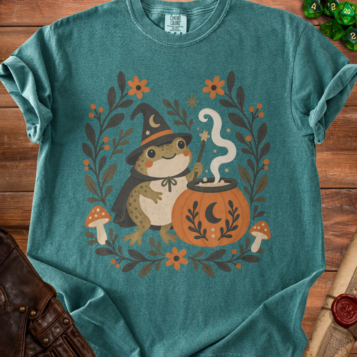 Frog wizard holding wand next to cauldron on teal t-shirt with floral and mushroom border.