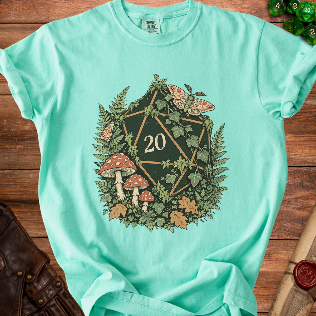 Teal t-shirt with a dark green twenty-sided die surrounded by ferns, mushrooms, and moths.