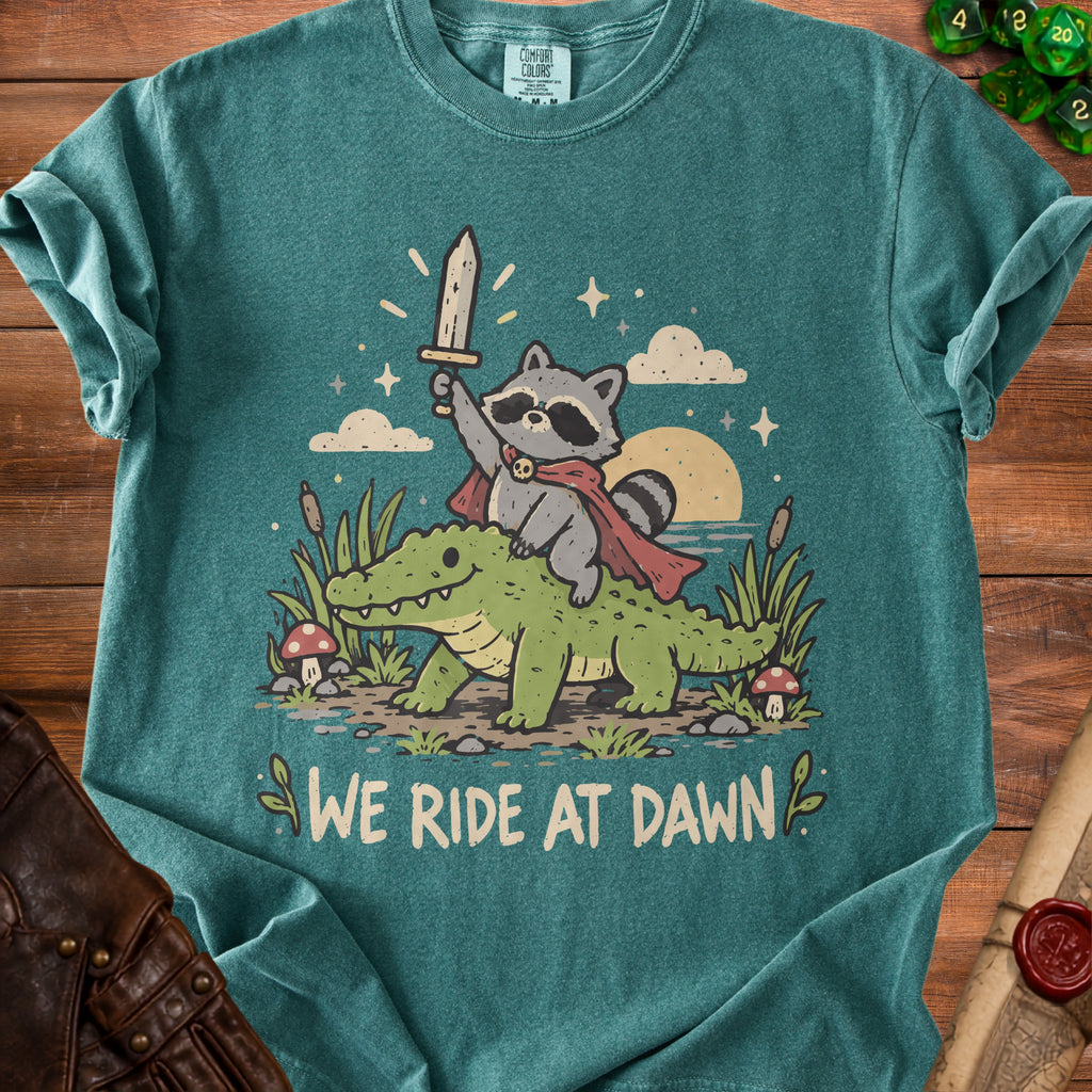T-shirt graphic: raccoon holding sword on alligator with "We Ride at Dawn" text.