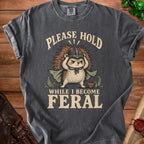 Gray t-shirt with illustration of a hedgehog in armor and text "PLEASE HOLD WHILE I BECOME FERAL".