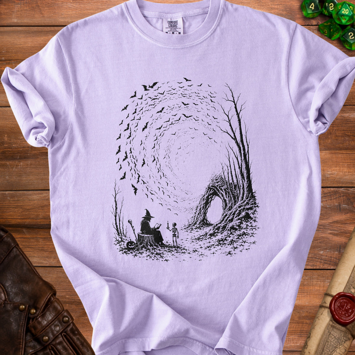 Light purple t-shirt with black graphic of a witch, skeleton, and bats.