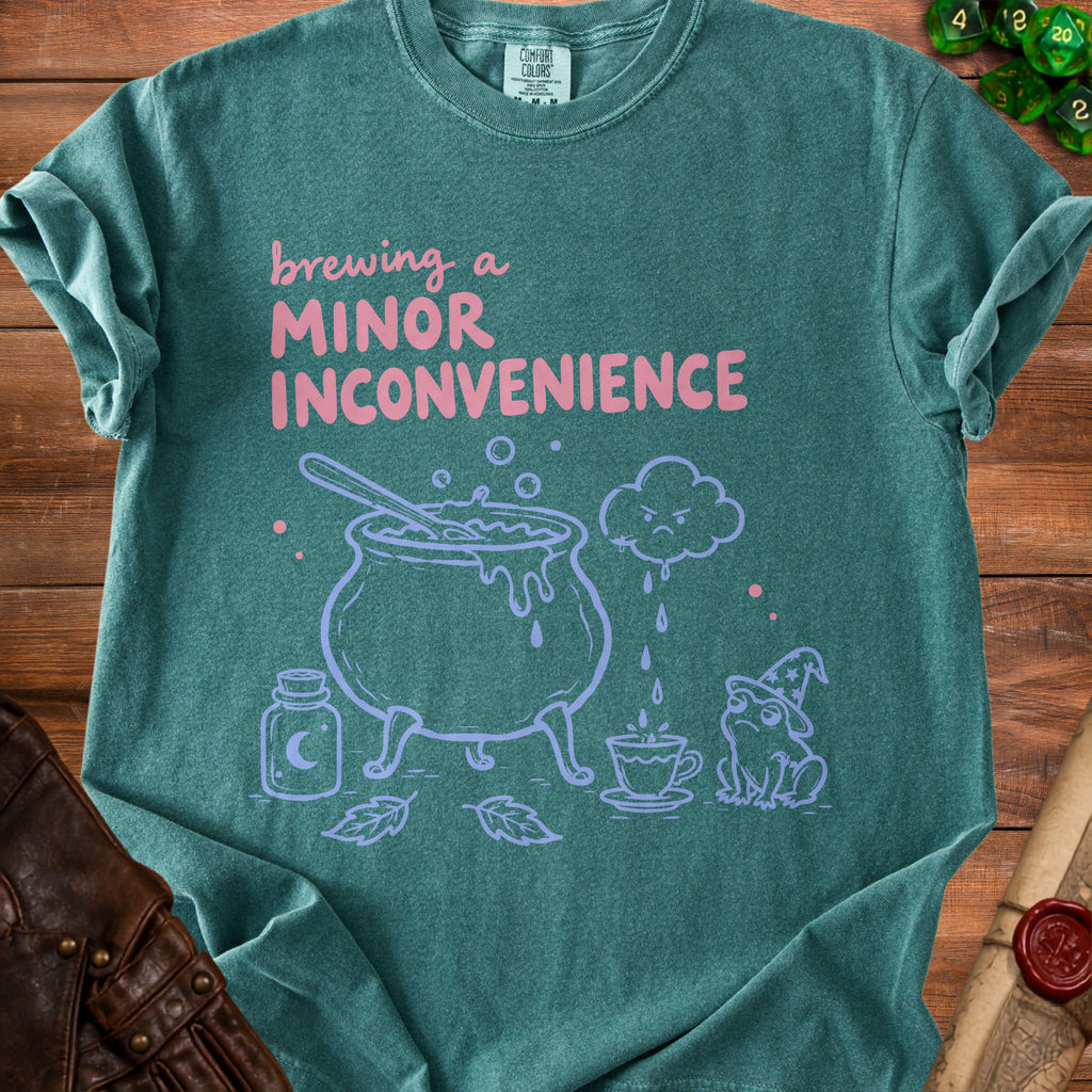 Teal t-shirt with "Brewing a Minor Inconvenience" text and cauldron, frog, and potion graphics.