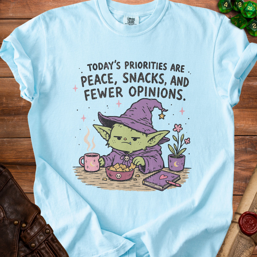 Light blue t-shirt with a goblin wearing a witch hat, eating snacks, and text about priorities.
