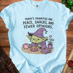 Light blue t-shirt with a goblin wearing a witch hat, eating snacks, and text about priorities.