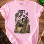Pink t-shirt graphic: skeleton rests with cat, text "Rest is a spell".